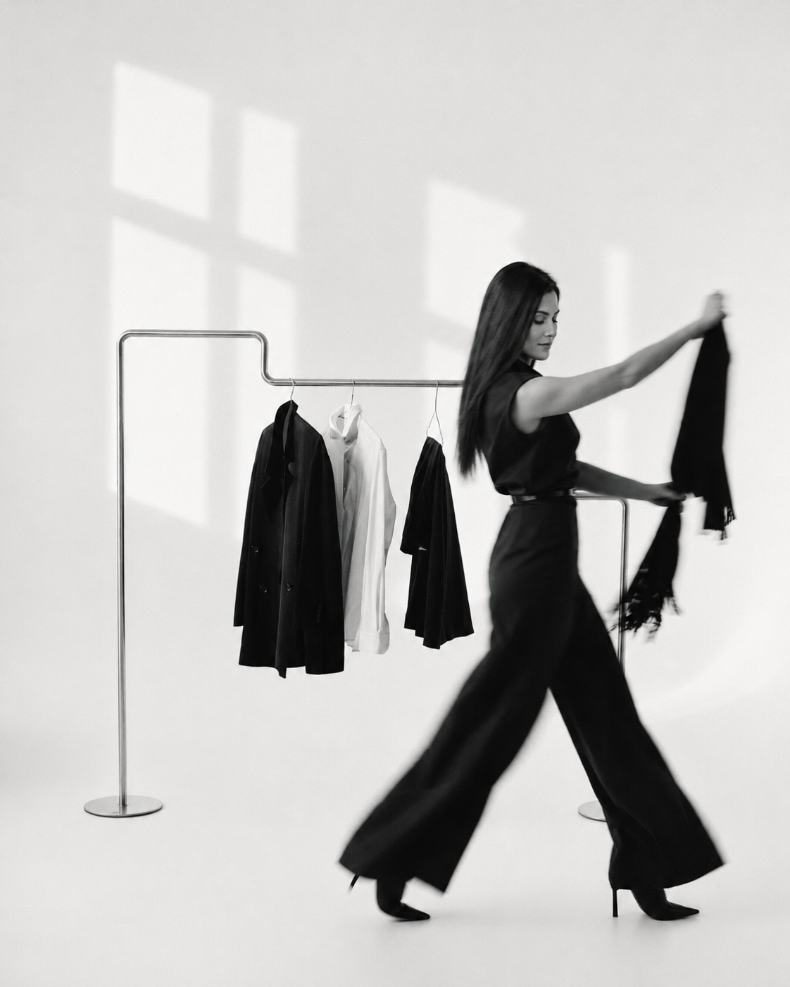 A woman in a black jumpsuit selecting garments from a clothing rail, an editorial black and white portrait.